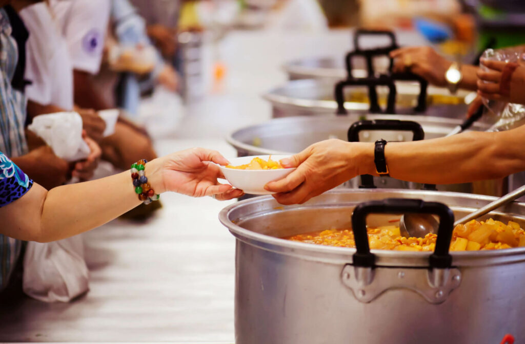 Volunteers serving food at a homeless shelter. the image is a close up of a large pot of soup, and a volunteer is handing a bowl full to a person standing in a line.