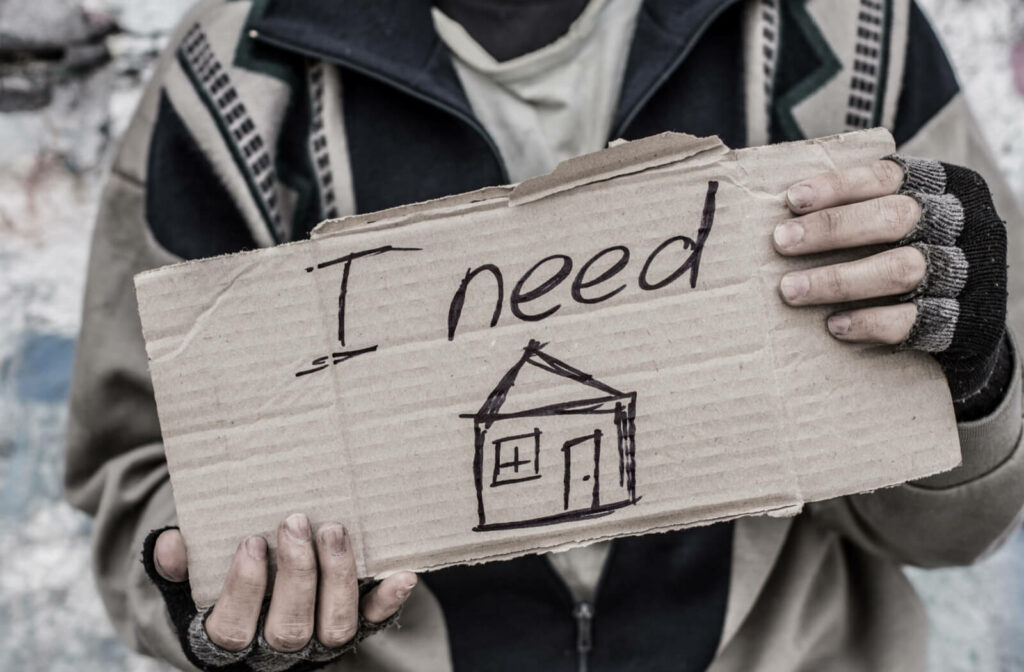 A man with dirty hands and clothes is holding a piece of cardboard with written words " I need a house".