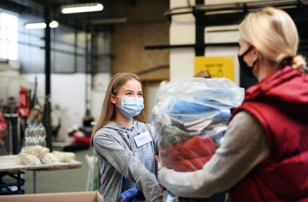A woman in a red bubble jacket is donating clothes received by a young girl in a donation receiving center.