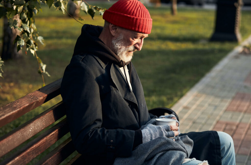 A senior man wearing winter clothes and sitting on a bench outside with a tin cup