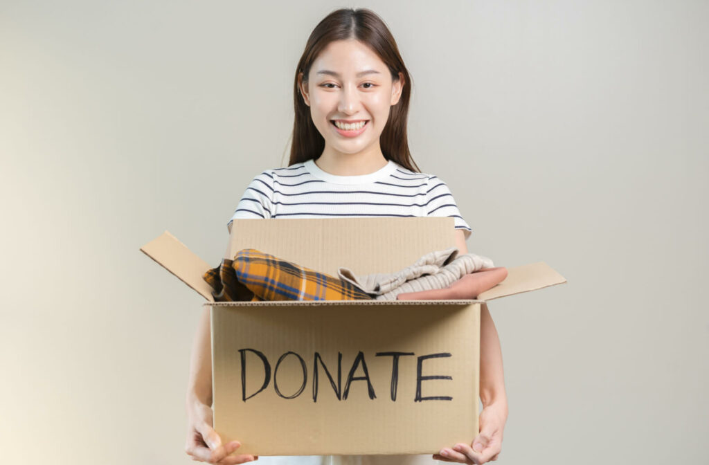 A woman in a striped shirt carrying a donation box filled with clothes against a beige background.