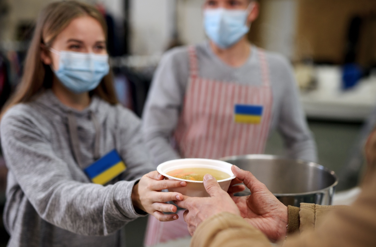 A volunteer in a soup kitchen with a mask on her face smiles while handing a bowl of soup to someone.
