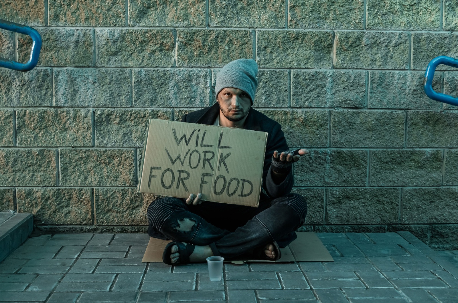 A person experiencing homelessness sitting on a piece of cardboard on the ground with an empty cup in front of him and a sign in his right hand that reads will work for food.