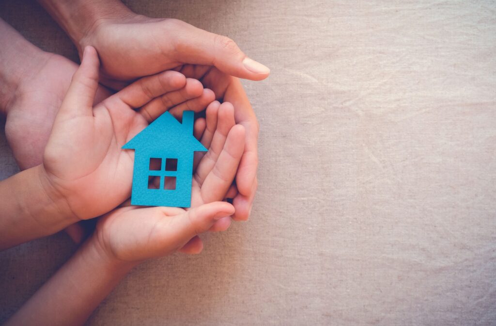 An adult's hands cradle those of a child, which in turn are holding a cut out of a blue house.