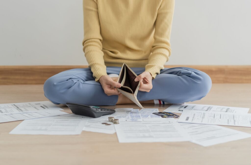 A person sitting cross-legged on the floor, surrounded by bills, is holding an empty wallet.