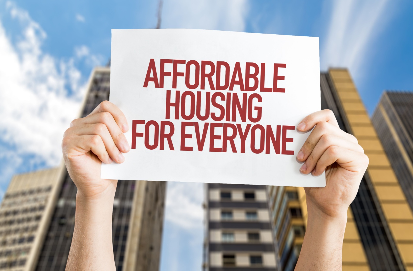 two hands holding a poster that reads, "Affordable housing for everyone" against a city backdrop.
