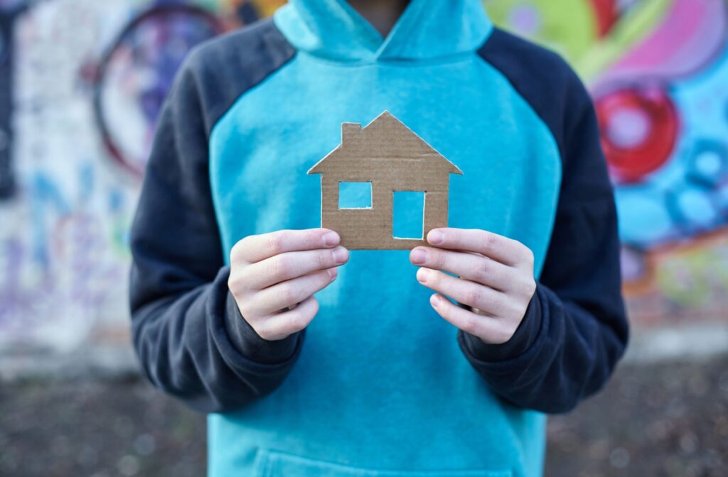 Close-up of a young boy in blue hoodie holding up cardboard cutout of a house.